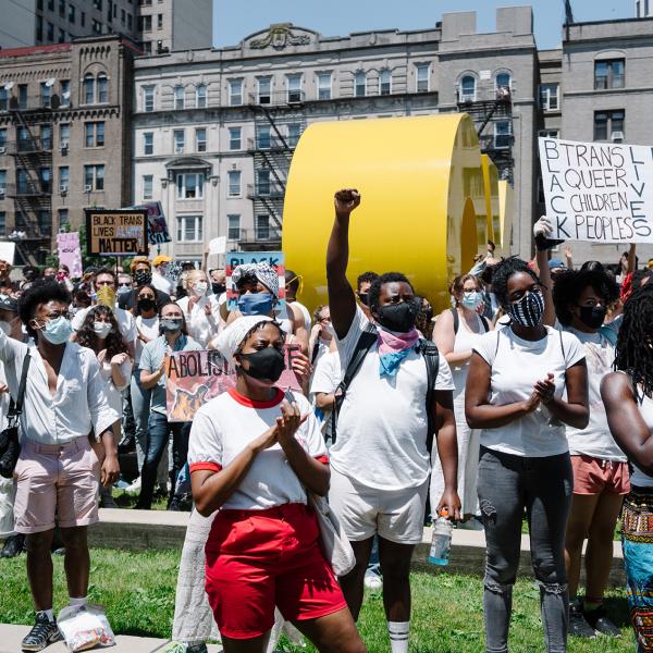 Una multitud de manifestantes que son mestizos pero predominantemente negros se solidarizan en la plaza frente al Museo de Brooklyn. Algunos llevan carteles, otros levantan los puños. Todos visten de blanco.