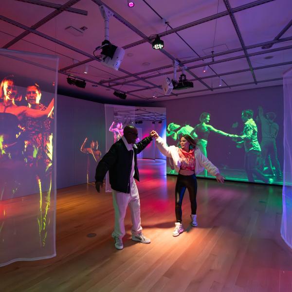 Two people dance together in a colorful, immersive art installation. The room features wooden floors and sheer fabric panels hanging from the ceiling, onto which vibrant, life-size video projections of dancers are displayed. The walls also show large-scale projections of people dancing, creating a lively, energetic atmosphere. Overhead lighting adds to the vivid, multicolored ambiance of the space.