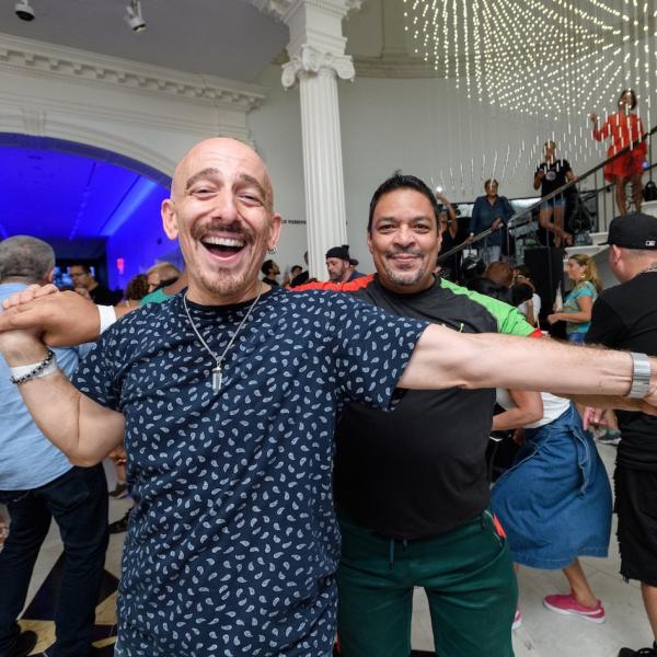 Two men smile and dance together in a room full of people.