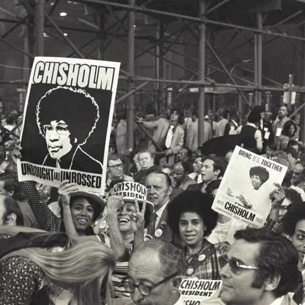 Uma fotografia em preto e branco de uma multidão reunida em apoio à candidata Shirley Chisholm. Várias pessoas estão sorrindo e segurando cartazes apoiando sua campanha para presidente.