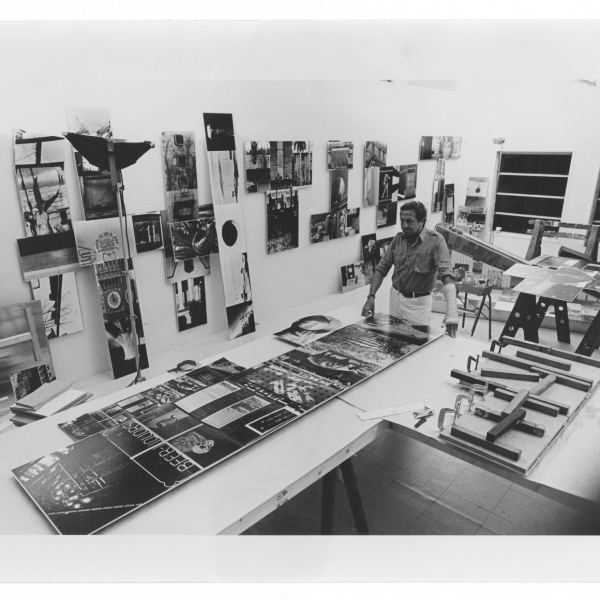 Rauschenberg stands in an art studio, examining large prints on a table surrounded by tools and materials, with artworks and photographs displayed on the walls. 