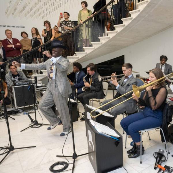 Dandy Wellington, a Black man in a gray striped suit dances in front of his seven piece jazz band in the Museum's rotunda. Behind them, guests in 1920s dress enjoy the performance. 