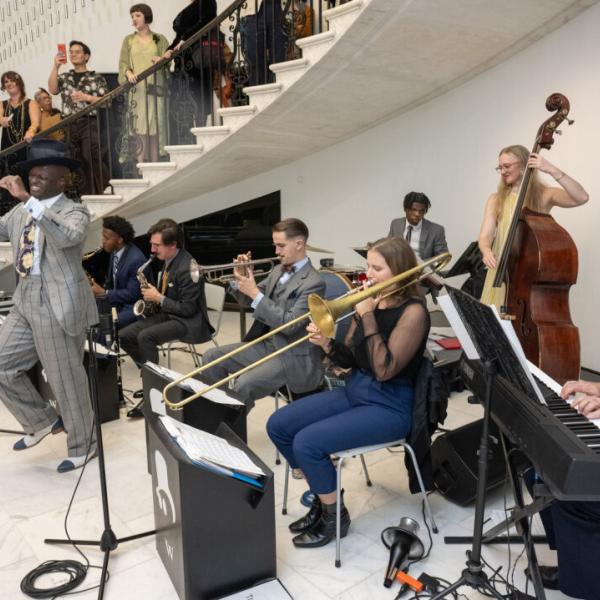 Dandy Wellington, a Black man in a gray striped suit dances in front of his seven piece jazz band in the Museum's rotunda. Behind them, guests in 1920s dress enjoy the performance. 