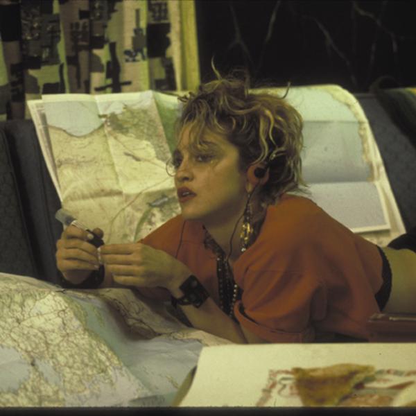 A still from Desperately Seeking Susan. Madonna is lying on her stomach on a couch while looking at a map and holding a pink telephone.