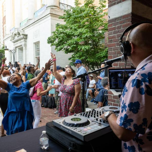 DJ Ted Smooth is at his turntables spinning in the foreground, facing away from the camera. Dancers in front of him throw their arms in the air.