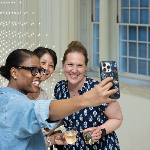 Three attendees smile for a selfie with drinks in the Museum’s Marble Court, with Starlight glowing in the background.