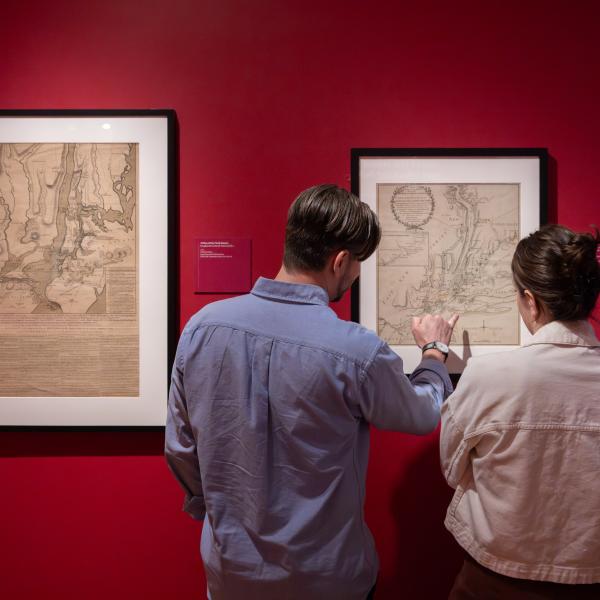 Two teachers view and discuss framed historic maps displayed on a red gallery wall in a museum exhibition.
