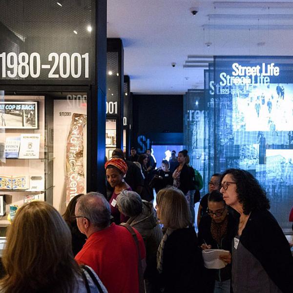 A group of educators gathered in the Museum of the City of New York’s “World City” gallery. Prominent displays include sections titled “1980–2001” and “Street Life,” with exhibits featuring themes such as policing, the events of 9/11, and urban life. The room is dimly lit with illuminated display cases and projected images. Attendees appear engaged, reading, discussing, and taking notes. 