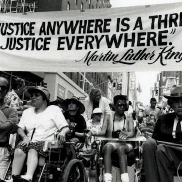 A crowd of people with disabilities and individuals in wheelchairs gather under a banner that reads "Injustice Anywhere is a Threat to Justice Everywhere" Martin Luther King Jr.