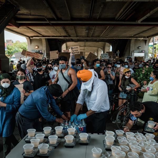 Una multitud de personas se paran alrededor de una mesa con recipientes de comida. El hombre en el centro está abriendo una caja de botellas de agua para distribuir.