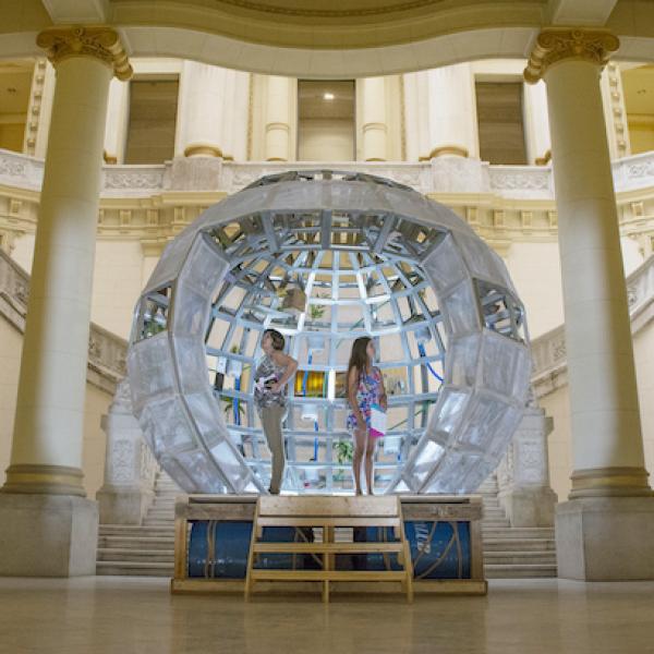 Two women stand in the center of a raised clear sphere, similar to a snow globe, set in a large open room with four white columns.
