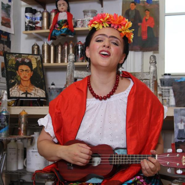 Foto de “The Colors of Frida” do Teatro SEA | Los colores de Frida ”apresentando a encenação da artista de Frida Kahlo em seu estúdio rodeada de pinturas e tocando um instrumento.