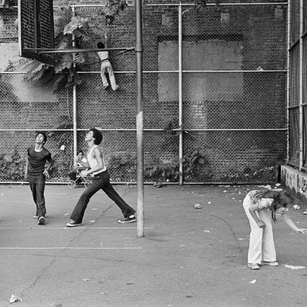 Photograph of people playing basketball in basketball court between two buildings, woman and child in front