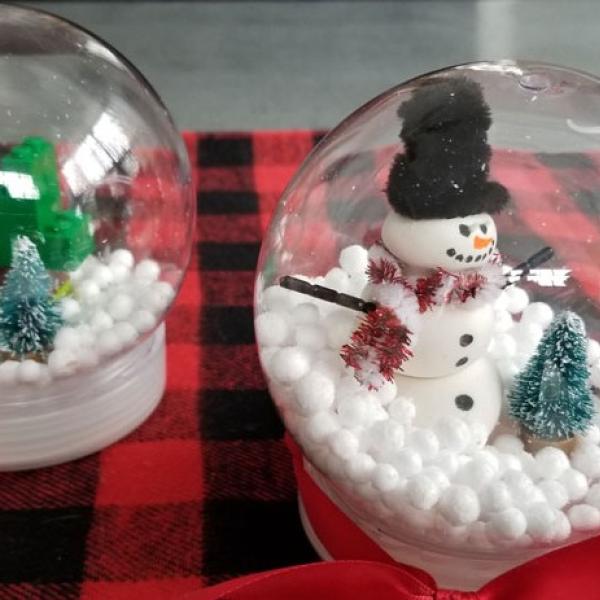 Image of two snow globe crafts. A winter scene with evergreen trees to the left, and a snowman and tree to the right.