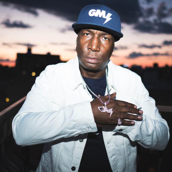 Grandmaster Flash is standing outside during sunset. He is wearing a dark blue baseball cap, a white jean jacket, and a black t-shirt. His right hand is holding up a gold chain with a pendant that has his initials, GM.