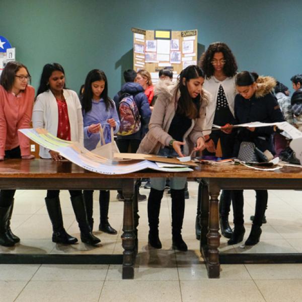 A group of students stand on one side of a long wooden table looking at documents and History Day projects.