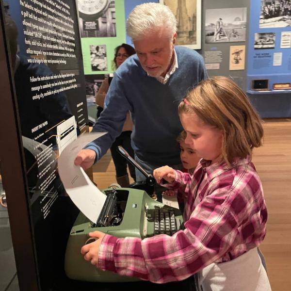 An older gentleman helping a young girl feed paper into a typewriter in the exhibition Analog City.