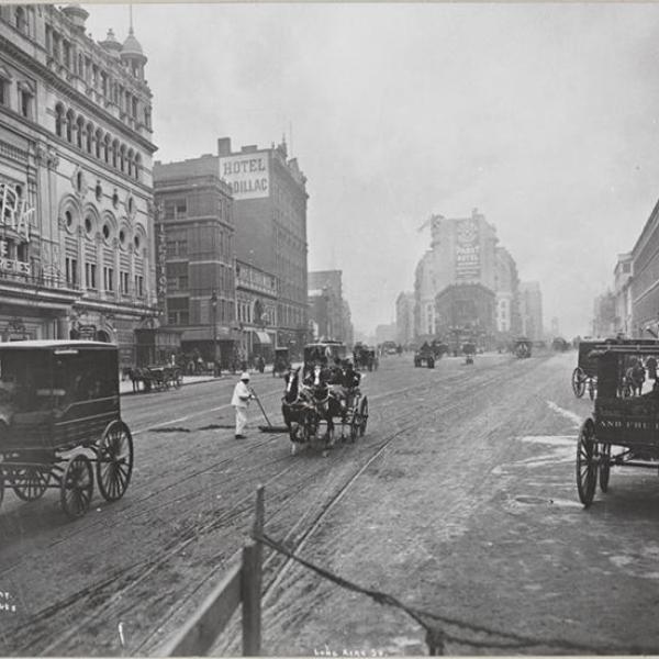 A black-and-white photograph showing the intersection of Broadway and 42nd Street, the present-day location of Times Square. The city street is surrounded by multistory buildings, and horse-drawn carriages drive in the street. In the center of the street is a New York City Sanitation worker dressed in white.