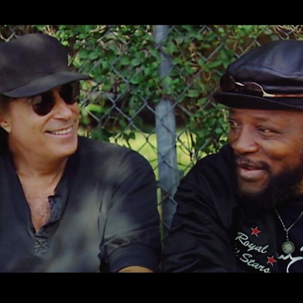 Two men sitting in front of a fence smiling 