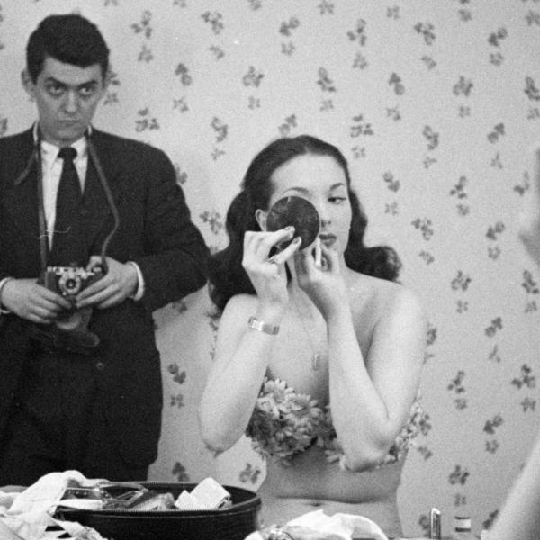 Taken in a mirror’s reflection, a woman uses a compact mirror to apply makeup, while photographer stands behind her watching