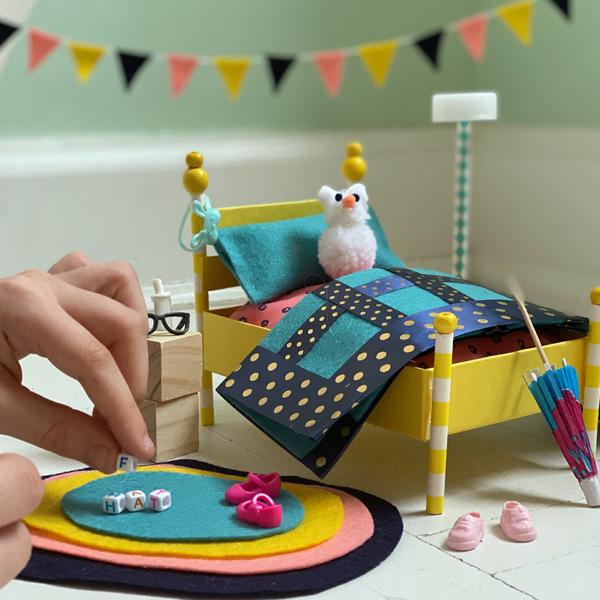 A photograph of a colorful set of miniature bedroom furniture, including a bed with stuffed animal, nightstand, rug, and decorations. An adult’s hand holding a tiny block is entering the view from the left, as if about to place it on the rug.