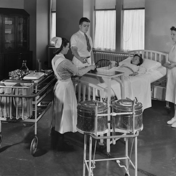 A black and white photograph of three nurses standing around a patient in a hospital bed.