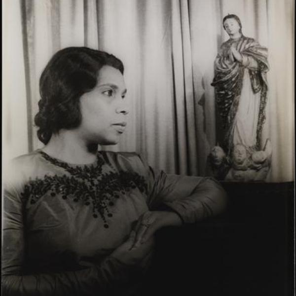 Black and white portrait of Marian Anderson in front of a curtain. Her arms rest on a dark surface, with a statue of a religious figure next to her left elbow.