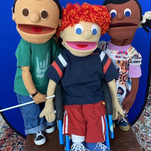 3 puppets created by NYC Kids Project standing on brown wood table, with bright blue background. One male puppet, Latinx, with dark hair, green tshirt , blue jeans , is holding a white cane. One male puppet with light skin and red hair uses a wheelchair and wears red pants, blue shirt with stripes on the arms. One female puppet, African-American has a colorful flowered dress over a lavender shirt and hair in two braids with white bows.  