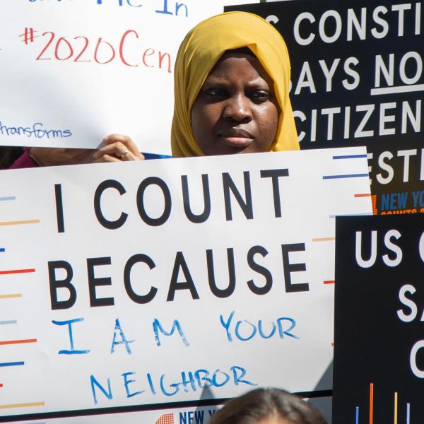 A imagem mostra um jovem em uma conferência de imprensa do Conselho da Cidade de Nova York segurando uma placa com a inscrição “Eu conto porque sou seu vizinho”.