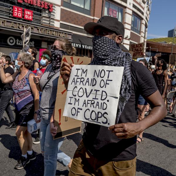 마스크를 쓰고 표지판을 들고있는 행진들의 군중. 한 사람은 "저는 COVID를 두려워하지 않습니다. 경찰이 두렵습니다."라고 말합니다.