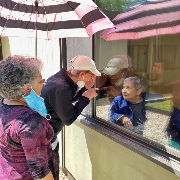 An older couple with their masks hanging on one ear talk to an older woman through a window.