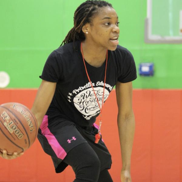 Priscilla Edwards holds a basketball in her right hand, in a low dribbling stance, she is facing a group off camera, speaking to them.