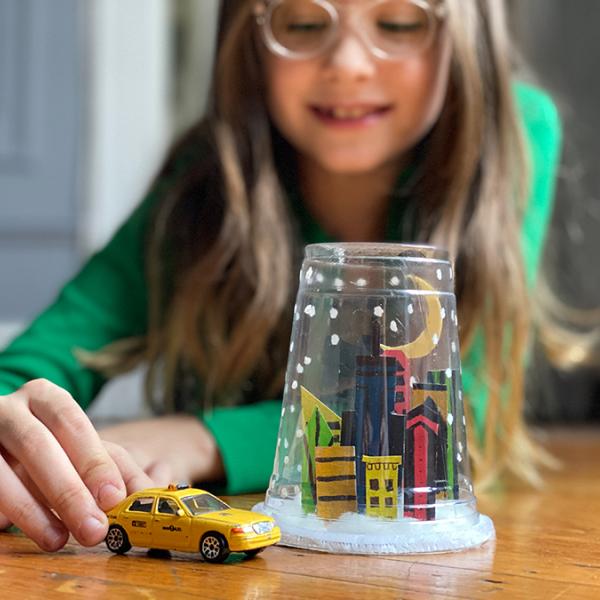 A young girl pushing a toy NYC taxi cab next to a snow globe made of a plastic cup with a hand-drawn city skyline  inside.