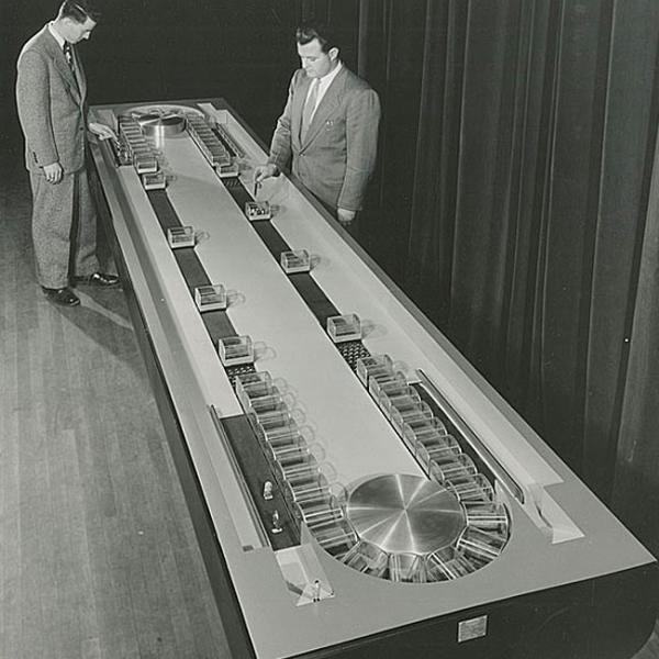 Two men in suits examining a working model of conveyor subway system.