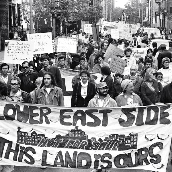 ローワー イースト サイドの通りを行進する大人のグループの白黒写真。 先頭にいる人々は「ロウアー・イースト・サイド。この土地は私たちのもの」と書かれた横断幕を掲げている。