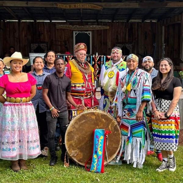 한 무리의 남성과 여성이 아메리카 원주민 전통 의상과 현대 의상을 혼합한 옷을 입고 눈에 띄게 전시된 드럼 주위에 서서 카메라를 향해 미소를 짓고 서 있습니다.