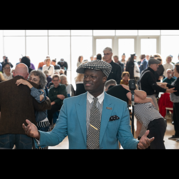 Performer Dandy Wellington dressed in a blue suit and patterned cap stands in front of a microphone, engaging with a lively crowd of people dancing and socializing in a bright, sunlit room