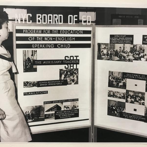 A woman stands in front of a Recruitment Board for Spanish-speaking Substitute Auxiliary Teachers in New York City public schools.  