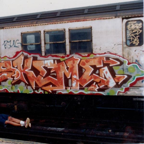 Skeme lies on the subway track below a parked subway car with a large orange artwork by him splashed across it. 