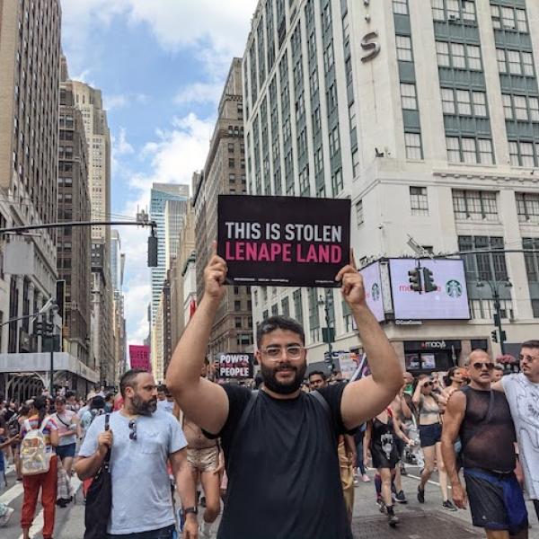 Un homme en marche dans une rue de la ville porte un petit panneau noir qui dit "Ceci est volé Lenape Land" en texte rose et blanc.