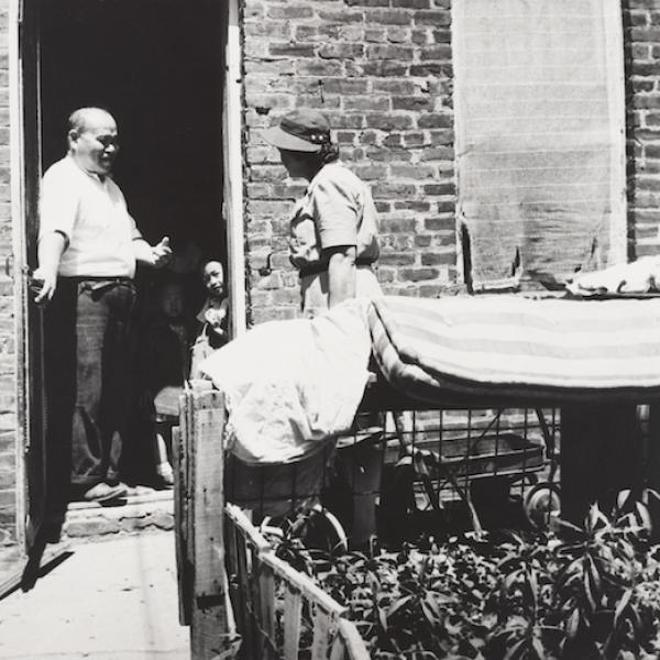 A man stands in a doorway with a small child. A woman is speaking to them from outside of a brick building.
