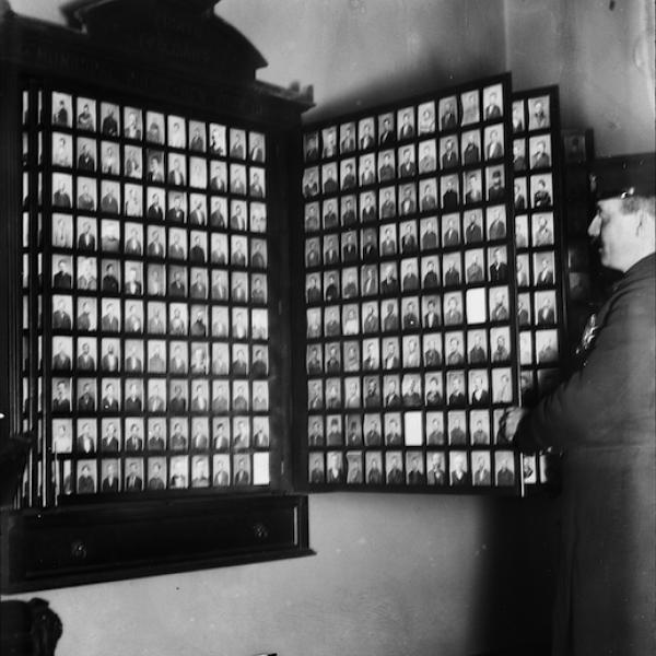 Man looking at a wall case with many tiny photographs of other individuals arranged in a grid.