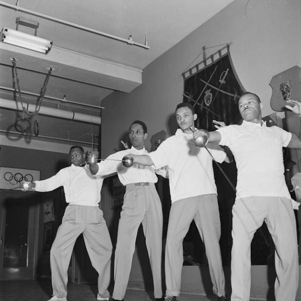 Foto preto e branco de quatro homens segurando sabres em pé em uma linha.