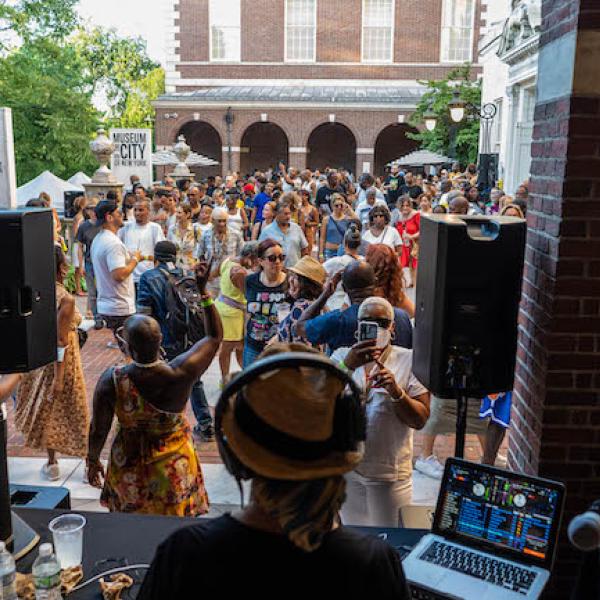 Vista de personas bailando en el patio del Museo en la entrada principal. El fotógrafo estaba parado justo detrás del DJ, por lo que la parte posterior de la cabeza del DJ está al frente. La gente habla, baila y bebe.