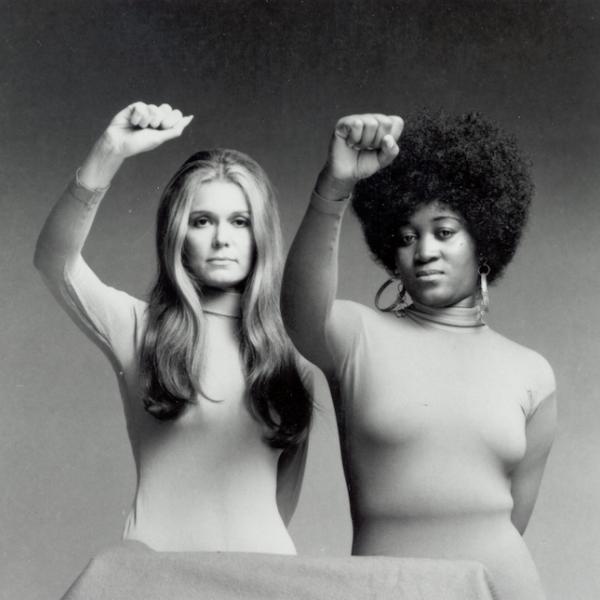 Goria Steinem and Dorothy Pittman Hughes stand together with their right hands raised in a fist above their heads