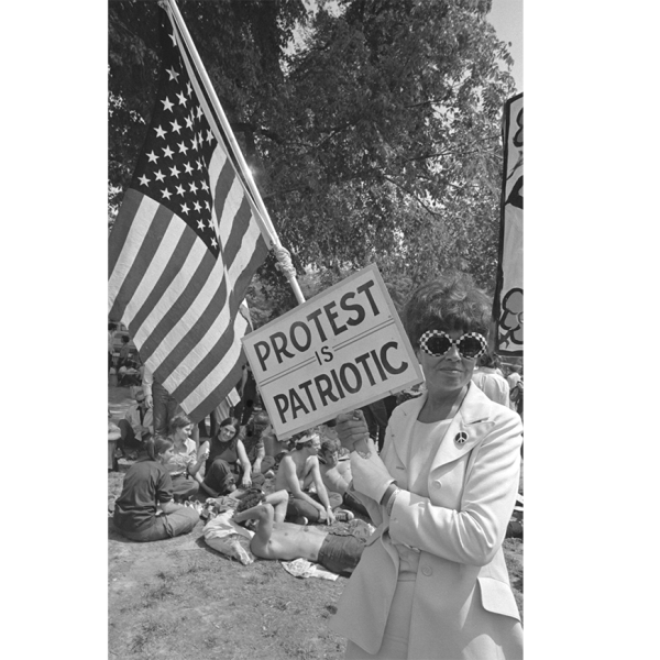 Uma fotografia do livro The Activist's Media Handbook, David Fenton. Uma mulher segura uma placa que diz "Protesto é patriótico" e a bandeira dos EUA.