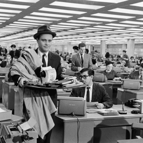 A still from the movie, The Apartment. A man is wearing a suit and holding office supplies, while walking through an office. 