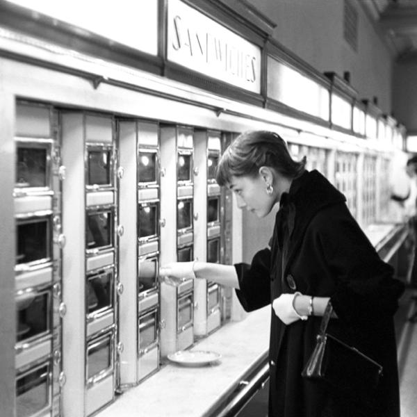 Audrey Hepburn is at an automat and opening a compartment to grab a sandwich. 