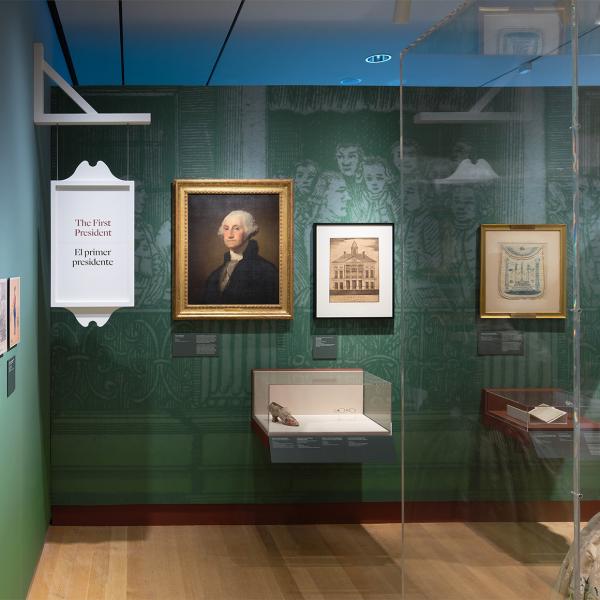 Installation view of the galleries, showing historic clothing, artwork on the walls, and a hanging sign saying, “The First President.”