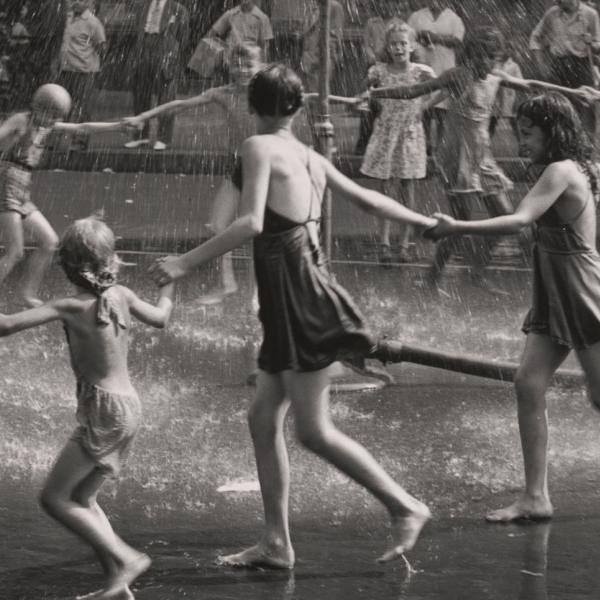 A group of children and teenagers hold hands in a circle as they run around a playground sprinkler